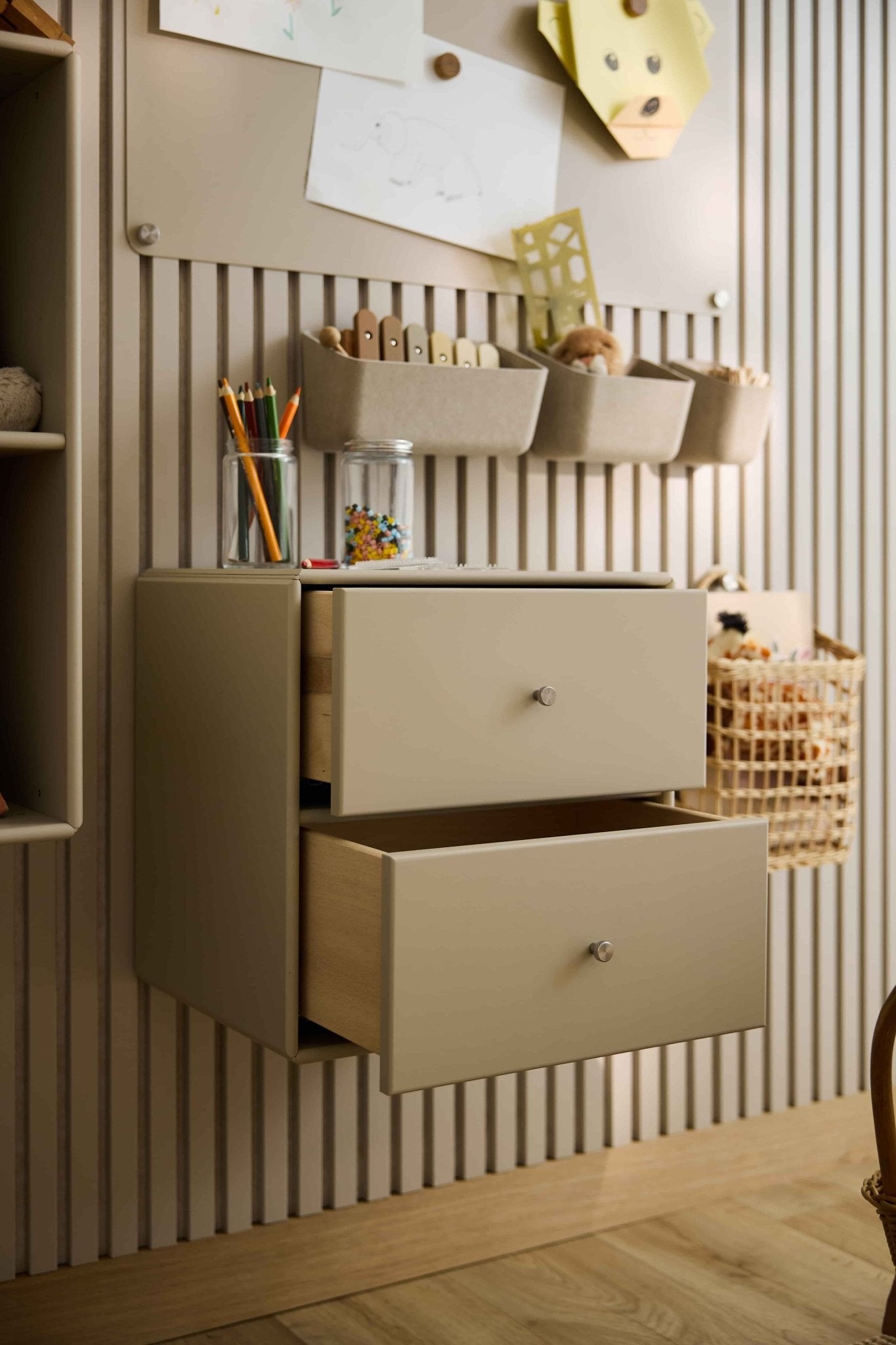 Dos cajoneras «Arka» de color beige de WoodUpp, parcialmente abiertas, sirven para guardar material artístico y juguetes; junto a ellas hay armarios cerrados y cestas.